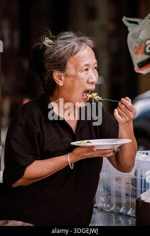 Bangkok, Thailand, 19. November 2025, eine ältere Frau, die auf einem geschäftigen Markt köstliches Street Food genießt. Stockfoto