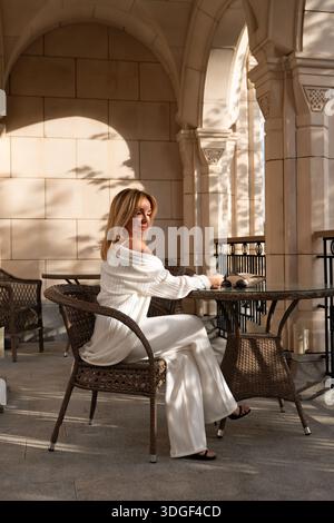 Frau White Outfit Patio Relaxing - Eine Frau in einem weißen Outfit entspannt sich auf einer Terrasse mit einem Tisch und Stühlen. Stockfoto