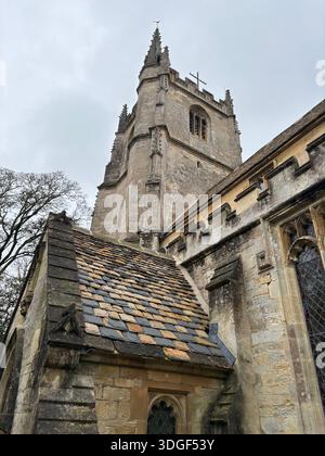 Historischer Kirchturm aus Stein, der an einem bewölkten Tag über einem traditionellen Schieferdach in einem Dorf der Cotswolds in England ragt. Stockfoto