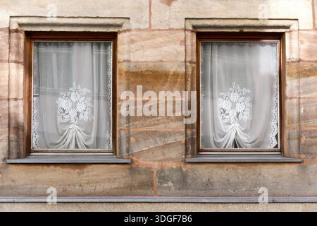 Zwei identische Fenster mit Spitzenvorhängen und Blumenmustern an einer historischen Steinfassade, die eine symmetrische und nostalgische Architektur schaffen Stockfoto