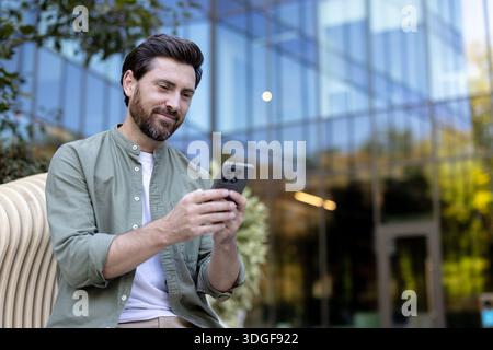 Mann mit Bart, der draußen sitzt, ein Smartphone hält und lächelt, reflektiert moderne Kommunikation, Technologie und Verbindung in einer ungezwungenen Geschäftsumgebung Stockfoto