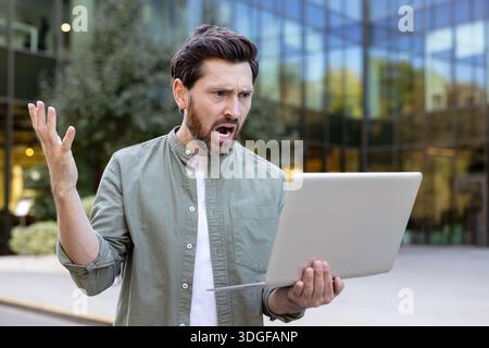 Ein junger, verärgerter Mann, der im Freien wütend aussieht und seinen Laptop anschreit. Er hat schwere Technologieprobleme, Probleme bei der Arbeit oder eine schlechte Internetverbindung, während er draußen arbeitet Stockfoto
