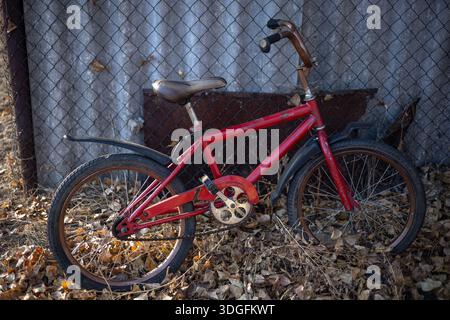 Ein rotes Kinderfahrrad mit braunem Sitz lehnt sich gegen einen rostigen Kettenzaun inmitten von herabfallenden Blättern. Stockfoto