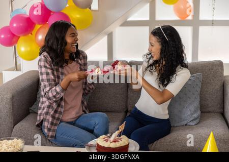 Diverse Mutter und Teenager-Tochter bieten im Wohnzimmer Kuchenscheiben mit Ballons an Stockfoto