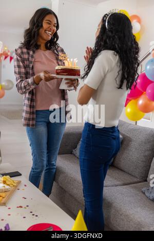 Vielfältige Mutter und Teenager-Tochter feiern Geburtstag im Wohnzimmer mit brennendem Kuchen und Ballons Stockfoto