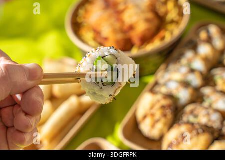 Sushi-Rolle mit Gurke, gehalten von Holzstäbchen vor einem bunten asiatischen Hintergrund zum Mitnehmen. Frischer Reis und Sesam sorgen für einen sauberen, appetitlichen Look Stockfoto