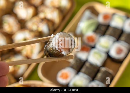 Lachs-Sushi-Brötchen mit Stäbchen vor einem gemischten Sushi-Tablett zum Mitnehmen. Frischer Fisch, Reis und Algen sorgen für ein klassisches japanisches Essen Stockfoto