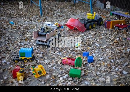 Bunte Plastikspielzeuge, darunter ein Lkw-Sockel, ein mehrteiliges Spielzeug und ein Aktivitäts-Spielzeug, sitzen in einem Sandkasten, der mit heruntergefallenen trockenen Pappelblättern bedeckt ist Stockfoto