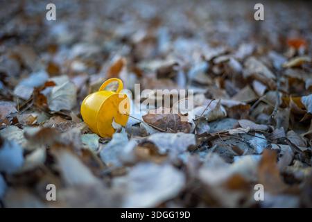 Ein kleiner gelber Spielzeugbecher liegt auf seiner Seite inmitten eines mit zahlreichen herbstlichen Pappelblättern bedeckten Bodens in Braun- und Beigetönen, unter Naturstein Stockfoto