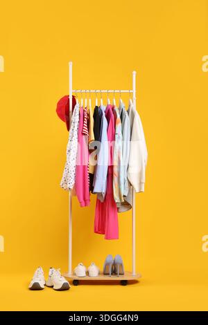 Verschiedene Kleidungsstücke hängen am Rack, Baseballmütze und Schuhe vor orangem Hintergrund Stockfoto