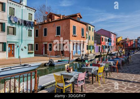 Die Insel Burano in der Lagune von Venedig, Italien. Restauranttische stehen für Gäste bereit an der Promenade am Kanal mit Blick auf traditionelle Häuser mit lackiertem fa Stockfoto