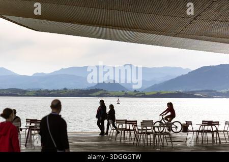 Santander, Spanien – 20. November 2021: Eine Frau mit rotem Kapuzenpullover fährt mit dem Fahrrad entlang der Uferpromenade in der Nähe von Cafés mit einem malerischen Berg V Stockfoto
