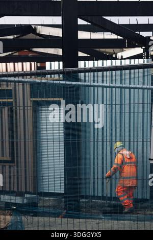 Bauarbeiter in gut sichtbarer orangefarbener Kleidung und Sicherheitshelm, der hinter einem Metallzaun auf einer Industriebau läuft. Stockfoto