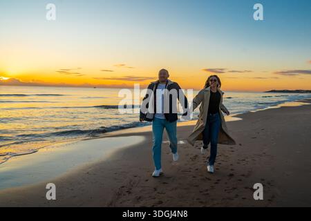 Strandspaziergang bei Sonnenuntergang, energiegeladene Paare spazieren an der Sandküste in der Abenddämmerung, Paar, die während des pulsierenden Sonnenuntergangs gezielt Schritte an der Küste machen Stockfoto