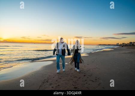 Menschen, die bei Sonnenuntergang gemütlich entlang der Küste spazieren gehen, Paar, die bei Sonnenuntergang Hand in Hand am Sandstrand entlang laufen Stockfoto