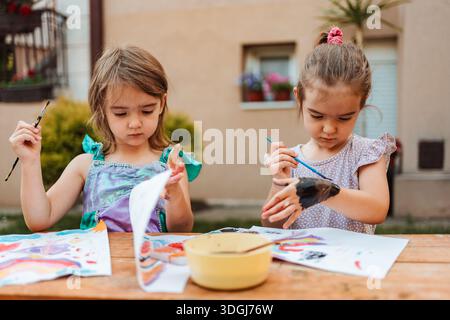 Zwei Vorschulmädchen malen mit Aquarellen auf einem Holztisch im Hinterhof Stockfoto