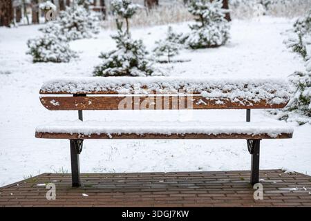 Eine hölzerne Parkbank ist nach einem Schneefall komplett mit frischem, weißem Schnee bedeckt. Die ruhige Winterszene unterstreicht die ruhige Schönheit einer einsamen Par Stockfoto