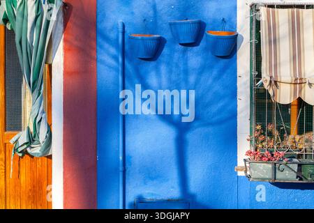 Drei blaue Töpfe und der Schatten eines Baumes bilden eine Komposition auf einer leuchtend blauen Wand neben einem Fenster mit Markise. Ein Konzept farbenfroher Straßendetails Stockfoto