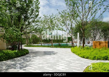 Ein friedlicher, gepflasterter Fußweg, der sich durch einen modernen Wohnpark oder Garten schlängelt. Der Weg ist von üppigen grünen Bäumen und gepflegten Landschaftsgärten umgeben Stockfoto
