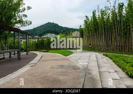 Ein friedlicher Parkweg, gesäumt von einem dichten Bambushain auf der einen Seite und einem hölzernen Sitzbereich auf der anderen Seite. Ferne Berge und Gebäude sind V Stockfoto