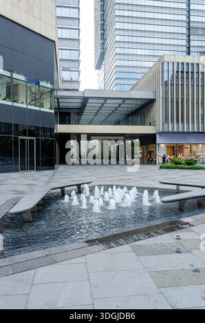 Vertikale Aufnahme eines kleinen Springbrunnens in einer modernen Fußgängerzone oder einem Einkaufszentrum plaza. Das Gebiet ist mit Stein gepflastert und umgeben Stockfoto