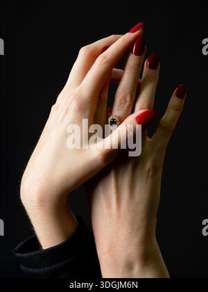 Studio-Aufnahme mit eleganten weiblichen Händen mit roten, gepflegten Nägeln und einem Ring, anmutig vor schwarzem Hintergrund, minimalistisches Schönheitskonzept. Stockfoto