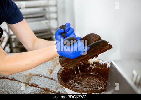 Ein handwerklicher Chocolatier, der blaue Nitrilhandschuhe trägt, kratzt geschmolzene dunkle Schokolade aus Spateln in eine Schüssel. Diese unordentliche Arbeit geschieht auf einem Granit C Stockfoto