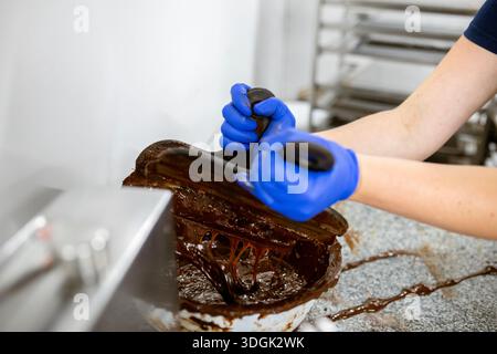 Ein handwerklicher Chocolatier, der blaue Nitrilhandschuhe trägt, kratzt geschmolzene dunkle Schokolade aus Spateln in eine Schüssel. Diese unordentliche Arbeit geschieht auf einem Granit C Stockfoto
