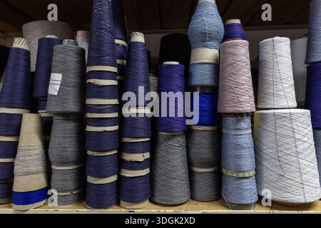 Nahaufnahme von Garnkonen in einem Regal in einem Strickwarenlager. Das Regal ist mit Spulen aus verschiedenen blauen, grauen und violetten Threas bestückt Stockfoto