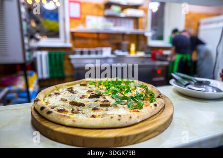 Köstliche heiße runde Pizza mit zwei verschiedenen Hälften auf einem Holzbrett steht auf der Theke in einer belebten Pizzeria-Küche mit Köchen, die im Unschärfe arbeiten Stockfoto