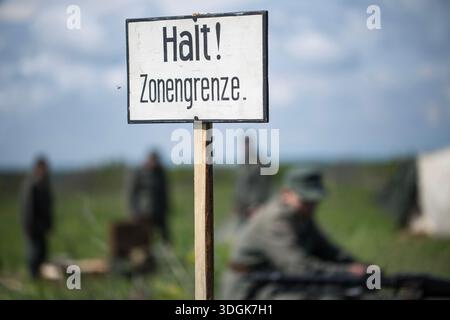 Weißes hölzernes Schild mit deutschem Text „Halt Zonengrenze“ bedeutet, dass Zone Border auf dem Feld in der Nähe von verschwommenen Soldaten in Vintage-Uniformen während einer Geschichte steht Stockfoto