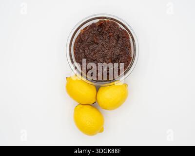 Eine minimalistische, hochwinklige Komposition mit einer indischen Zitronenpickle (Achaar) in einer Glasschale mit drei Reifen gelben Zitronen darunter. Stockfoto