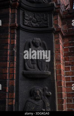 Dunkles Steinrelief auf einer roten Backsteinkirche in Kattowitz zeigt einen Engel mit einem Kreuzschild über einer gekleideten Figur, mit floralen Motiven, harkendem Frühlingslicht, Stockfoto
