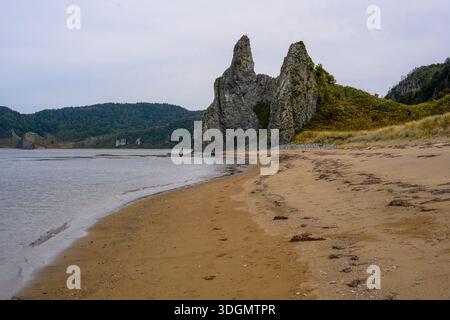 Die Wellen schlängeln sanft am Sandstrand, während einzigartige felsige Klippen vor dem Hintergrund grüner Hügel hoch stehen. Eine friedliche Szene fängt die Ruhe ein Stockfoto