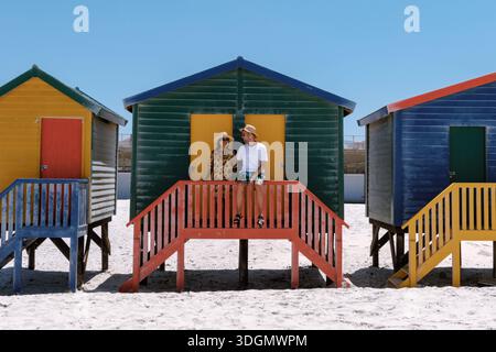 Zwei fröhliche Touristen stehen auf den Stufen der farbenfrohen Strandhütten am Muizenberg Beach. Kapstadt Südafrika Stockfoto