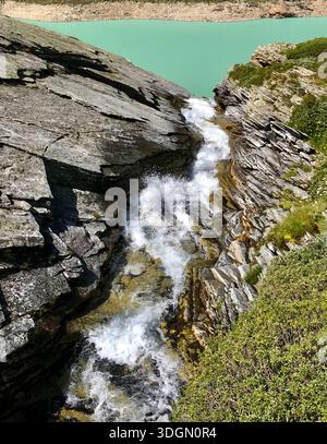 Zwischen zwei schmalen Felsspalten fließt ein beeindruckender Gebirgsbach, der sich in Richtung eines beeindruckenden Ausblicks auf den türkisblauen Mattmark See im Wallis, Schweiz, öffnet Stockfoto
