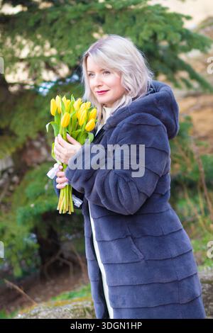 Eine nachdenkliche blonde Frau in einem Kapuzenmantel hält draußen gelbe Tulpen nahe an ihr Gesicht. Sanfter grüner Hintergrund, ruhiges Frühlingsporträt. Stockfoto