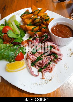 Eine weiße Platte mit Garnelen, Tomaten, Salat, Fischkuchen, einem Zitronenkeil, und Soße in einem weißen Becher sitzt auf dem Tisch. Die Garnelen und Fischkuchen sind Top Stockfoto