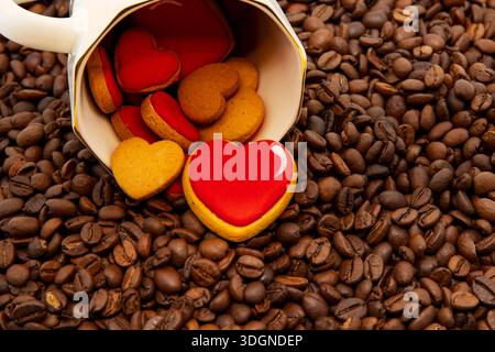Herzförmige Kekse, die aus der Kaffeetasse auf geröstetem Kaffeebohnenhintergrund verschüttet werden Stockfoto