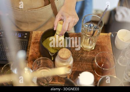 Professioneller Barista, der organische Matcha-Grünteepulver in einer Schüssel auf einem Holztisch mit Fokus und Hingabe an authentische japanische Be von Hand rührt Stockfoto