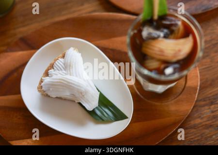 Köstliche Scheibe frischer Kokoskuchen mit cremeweißem Topping auf grünem Bananenblatt neben erfrischendem Eiskaffee auf Holztablett Stockfoto