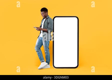 Junger Mann, der neben einem großen, leeren Smartphone steht, während er ein Gerät in hellem Raum benutzt Stockfoto