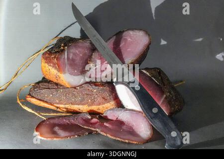 Rauchfleisch ist eine allgemeine Bezeichnung für Fleisch, das durch Poekeln und Kaltraeuchern haltbar gemacht wird. Sehr lecker in feinen Scheiben geschnitten auf einem Butterbrot mit Gewuerzgurken und Radieschen. Gehoert auf jedem Vesperbrett. Dazu ein kuehles Bier oder ein Glas Wein einfach herrlich auch sehr passend zu Spargel mit Kartoffeln. Zartes Rauchfleisch oder auch geraeucherter Lachsschinken direkt aus dem Raeucherofen, am Stueck und aufgeschnitten mit Messer - Rauchfleisch, Speck Rauchfleisch ist eine allgemeine Bezeichnung für Fleisch, das durch Poekeln und Kaltraeuchern haltbar ge Stockfoto