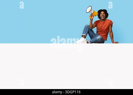 Positiver afroamerikanischer Mann, der auf leerem Brett sitzt und eine Ankündigung macht Stockfoto