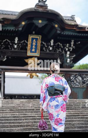 Frau in Yukata, die eine Steintreppe zum historischen hölzernen Tempeltor hinaufgeht. Kyoto, Japan - Juni Stockfoto