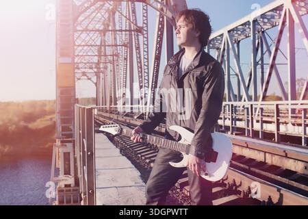 Ein Mann mit einer E-Gitarre auf der Eisenbahn. Ein Musiker in einer Lederjacke mit Gitarre auf der Straße im Industriegebiet. Gitarrist auf der Brid Stockfoto