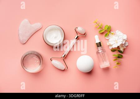 Jade-Gesichtsrollen und Gua-Sha-Steine für Beauty-Gesichtsmassage-Therapie, Artikel für die häusliche Behandlung mit Skin Care Anti-Aging-Tools, Gesichtsserum und Feuchtigkeitscreme auf rosa Hintergrund Stockfoto