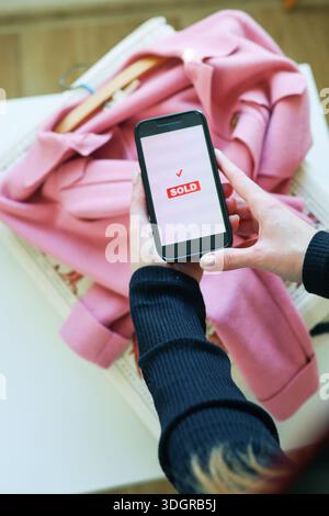 Smartphone mit Verkaufsbestätigung auf dem Bildschirm über pinkfarbener Kleidung. Online-Shopping, erfolgreicher Verkauf und Second-Hand-Modekonzept. Stockfoto