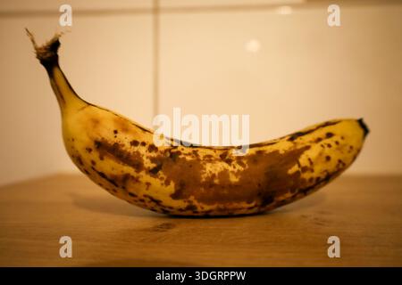 Eine perfekt gereifte Banane mit braunen Flecken liegt auf einer warmen Holzarbeitsfläche. Er leuchtet unter weichem Umgebungslicht und vermittelt ein Gefühl von Komfort und Einfachheit Stockfoto