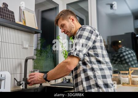 Mann, der am Spülbecken steht und Glas mit Wasser aus dem Wasserhahn füllt. Das Konzept des täglichen Routineverfahrens Stockfoto